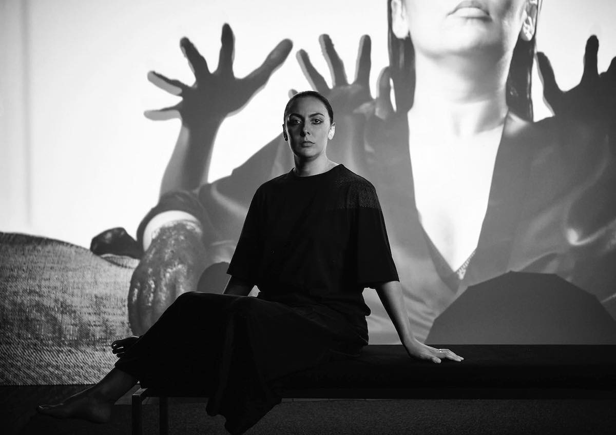 Woman wearing black sitting on a on a chair in front of a video projection of a woman with black hands reaching from behind her.
