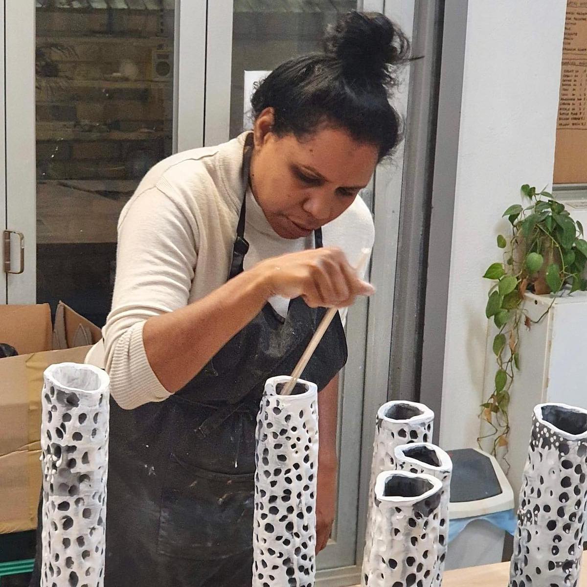 Image of a person wearing an apron as they hand paint the inside of a white and black ceramic sculpture, with other painted black and white ceramic sculptures nearby.