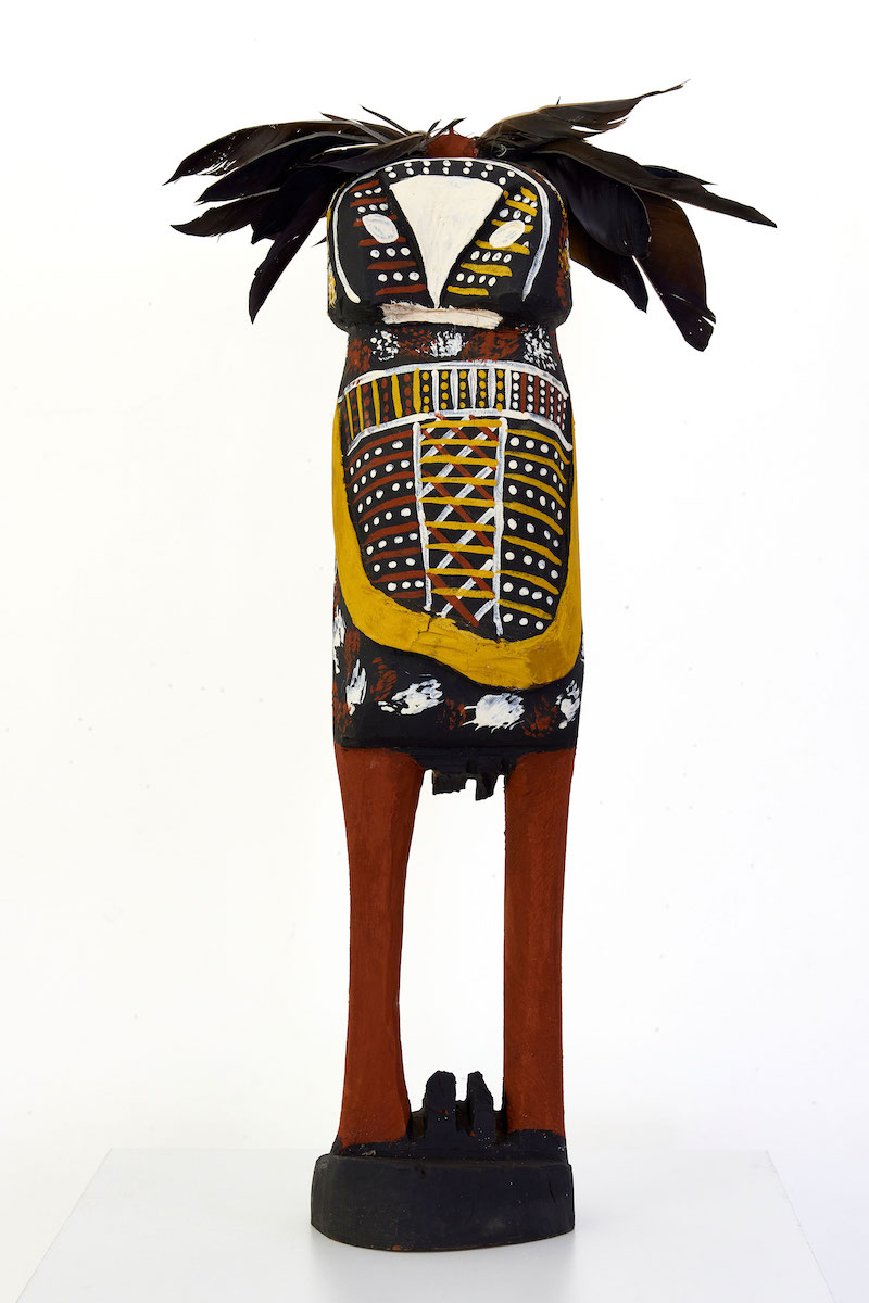 Carved, bird-faced figurative sculpture with head feathers and painted markings in black, red, yellow ochre and white.