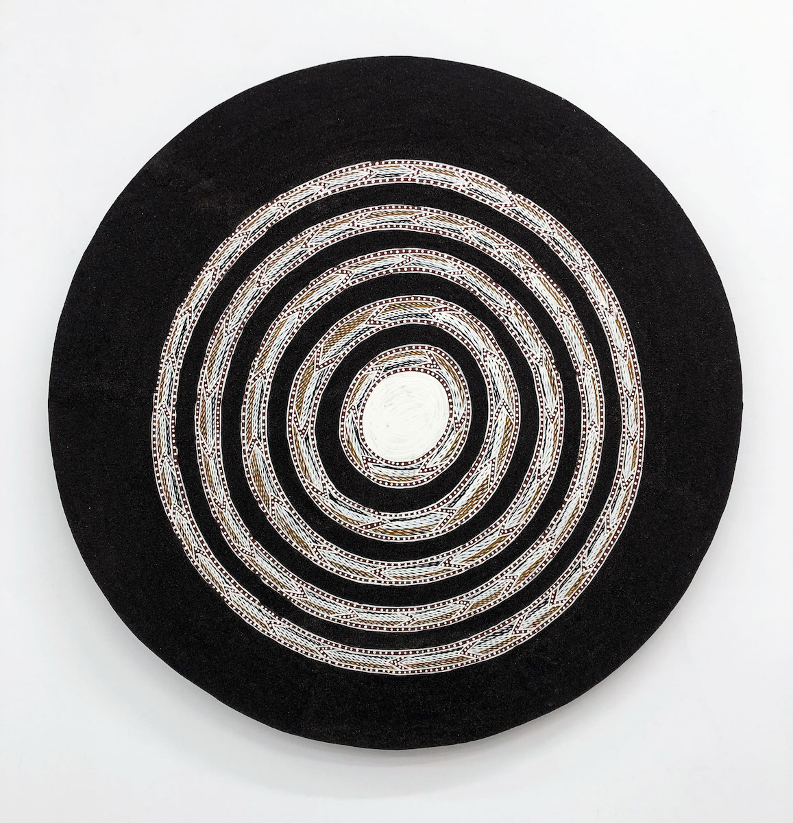 A circular board covered in shimmering black sand, with a painted white circle at its centre and concentric circles of geometric lines and dots in red, yellow and white emanating outwards