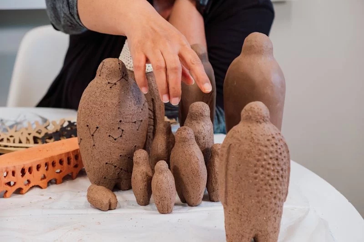 Image of a hand hovering above multiple brown clay sculptures in abstracted human form, pointing at another abstract clay human shape.