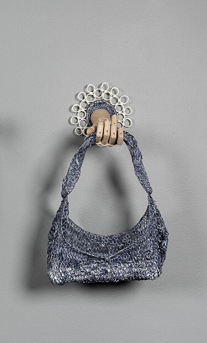 Blue-grey woven bag held by a wooden mannequin forearm protruding from the wall, framed by a series of white circles.