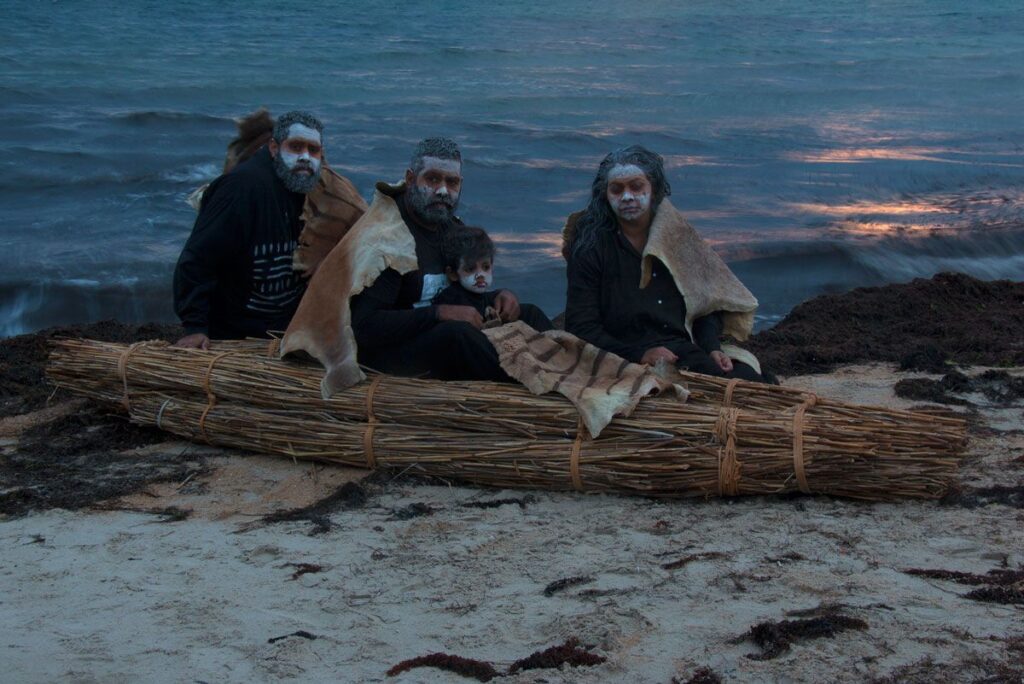 Three individuals in hoodies and animal skin cloaks sitting in a river-reed canoe on a sandy shore with the sea in the background.