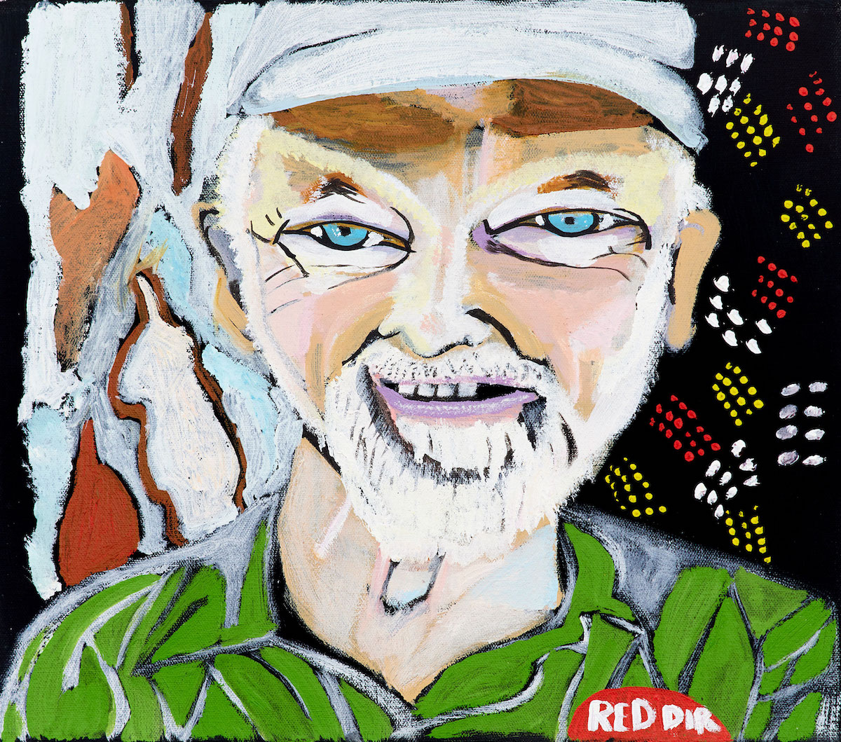 Portrait of a bearded person wearing a cap and a green striped shirt with a red sticker saying 'Red Dir' against an abstract, multicoloured background