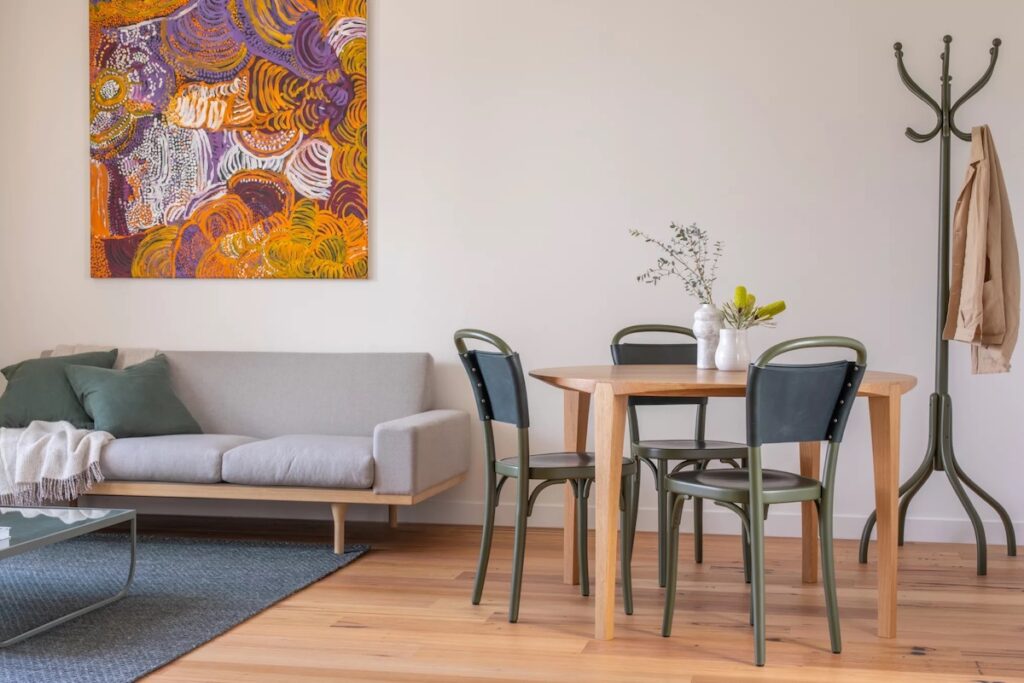 Image of a sleek hotel room with a couch, small dining table and chairs, a coat rack and a bright orange, purple and white Indigenous artwork.