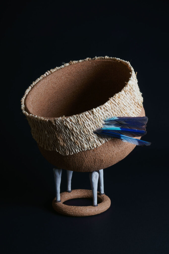 Photograph of an abstract sculpture in the shape of a bowl made from rough earth-coloured clay. The bowl is suspended in the air by four bone shaped pieces of white clay, upon a ring of earth-coloured clay. The rim of the bowl is clad in woven raffia, with two blue-coloured feathers attached to the side. Sculpture sits on a black background.