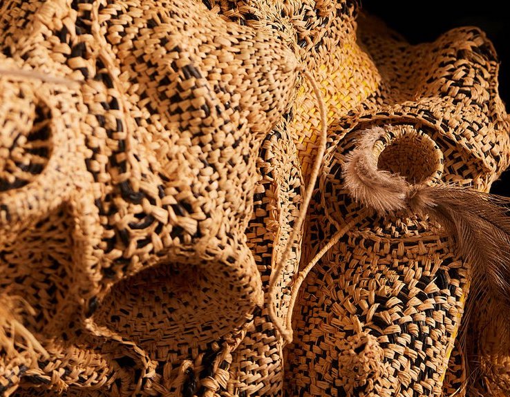 Close-up of an organically-shaped woven artwork with black, yellow and beige raffia and feather details.