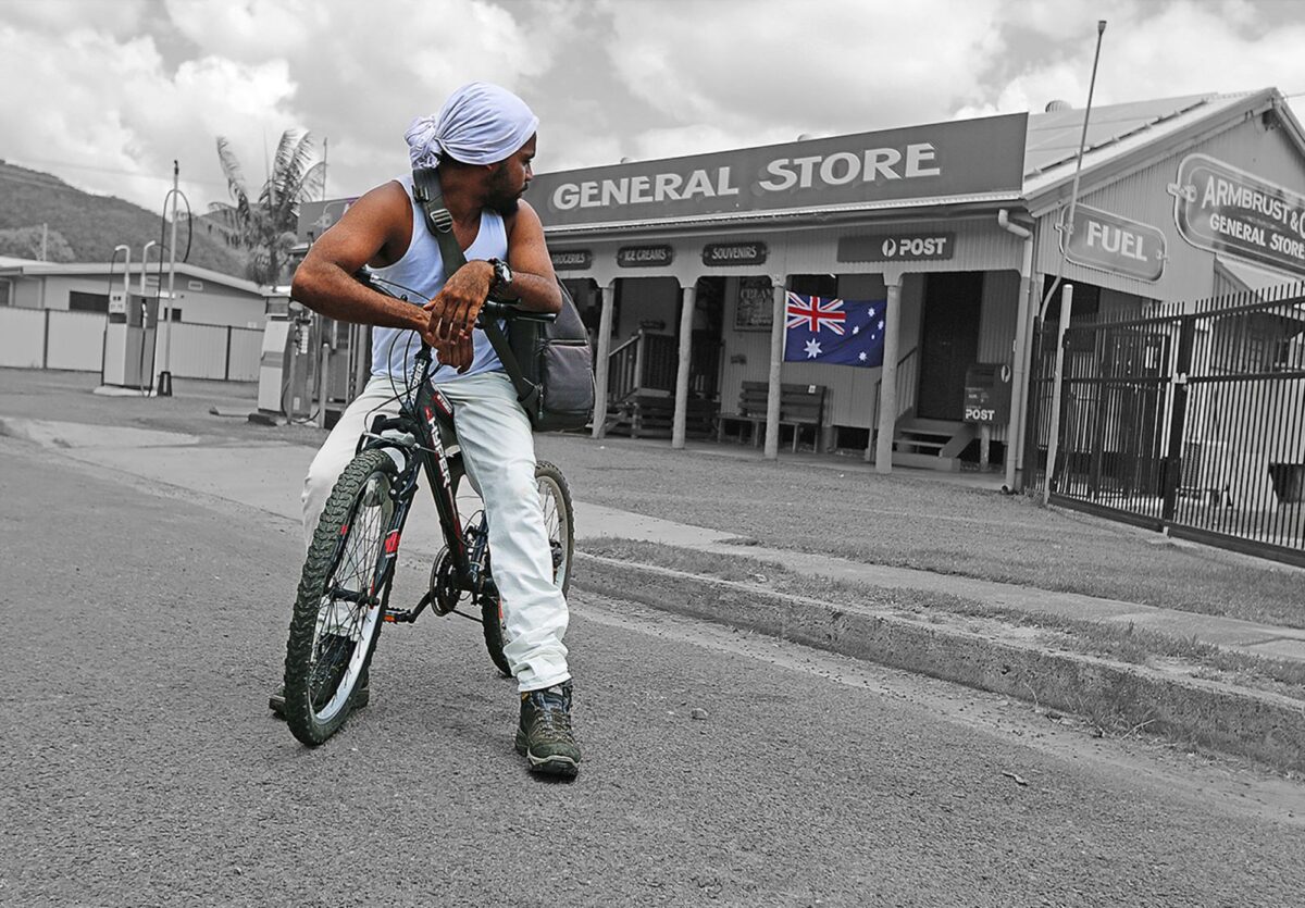 Young person on a bike looking back at the local general store from the road.