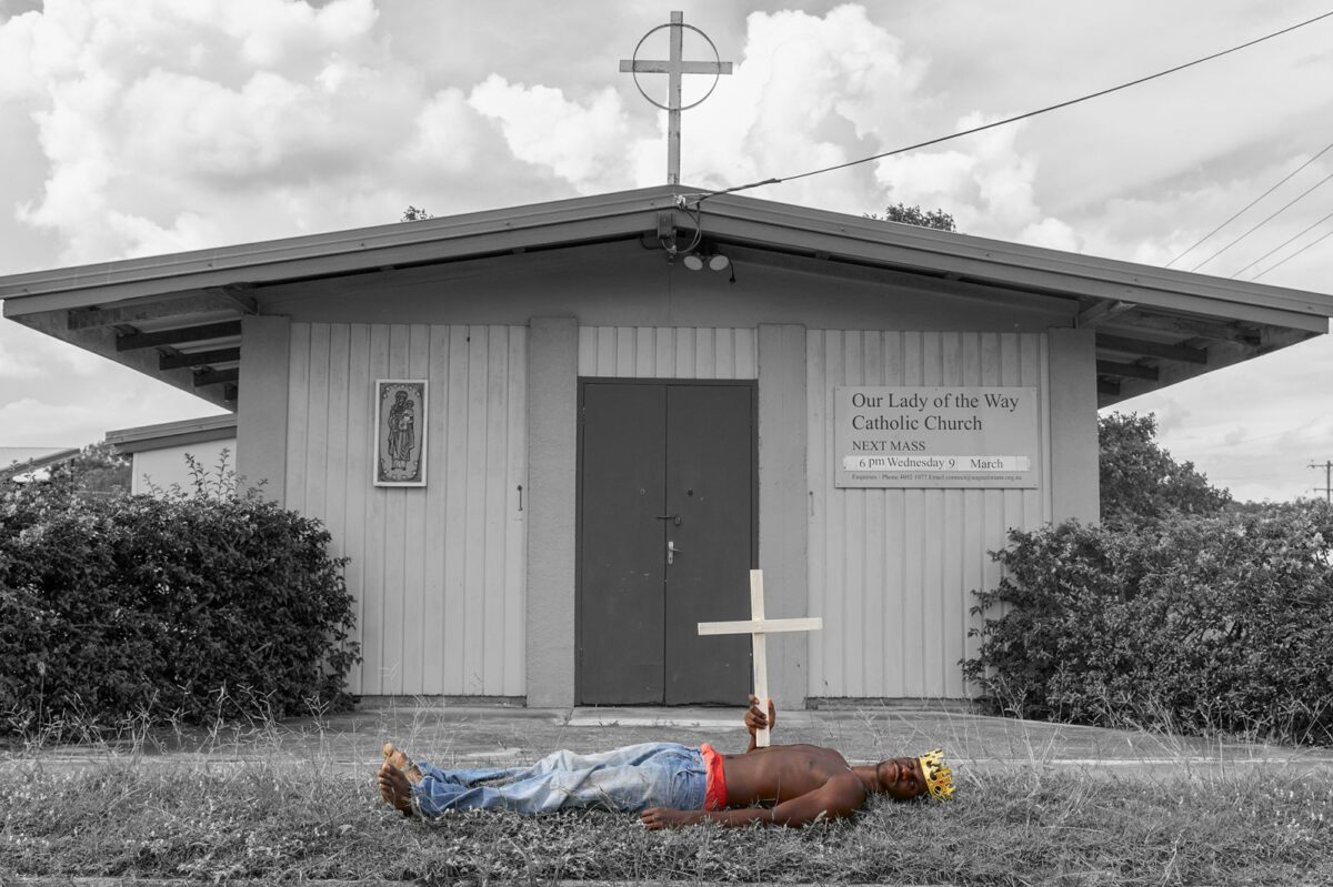 Young person laying on the ground in front of a church, holding a cross.