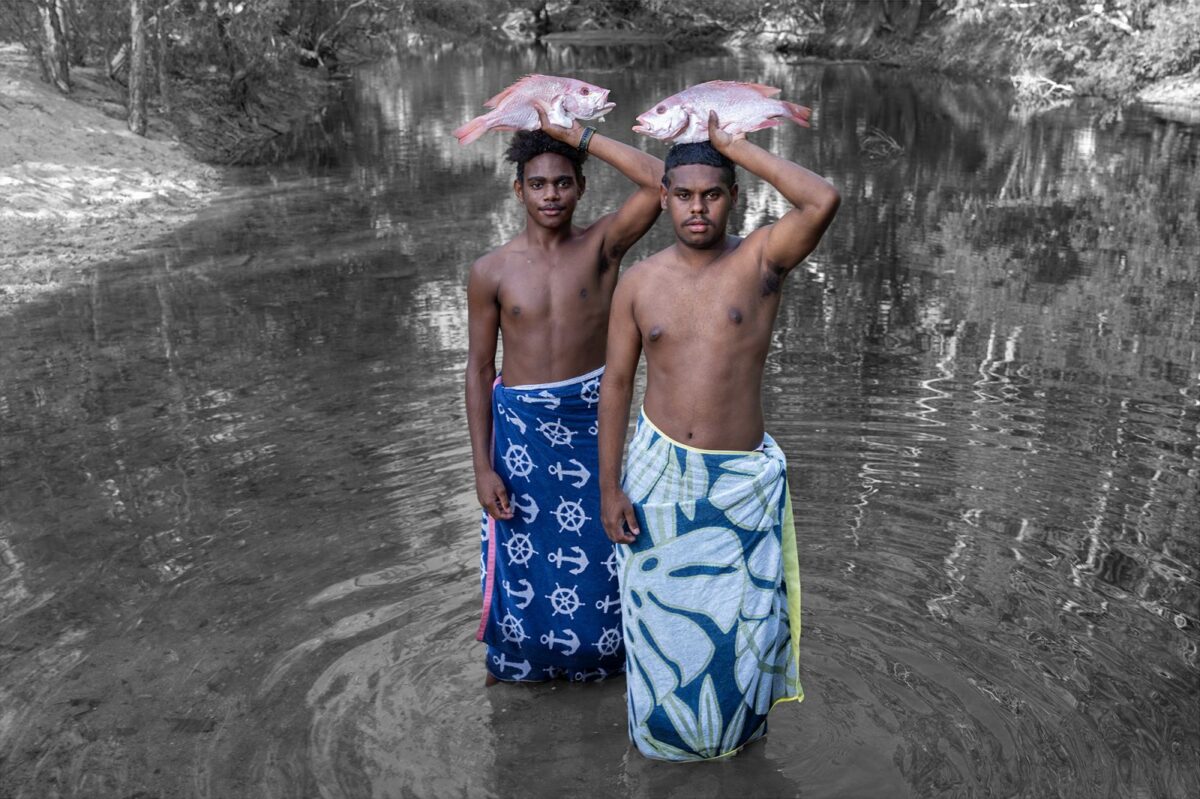 Two young people in towels standing in a body of water, holding fish over their heads.
