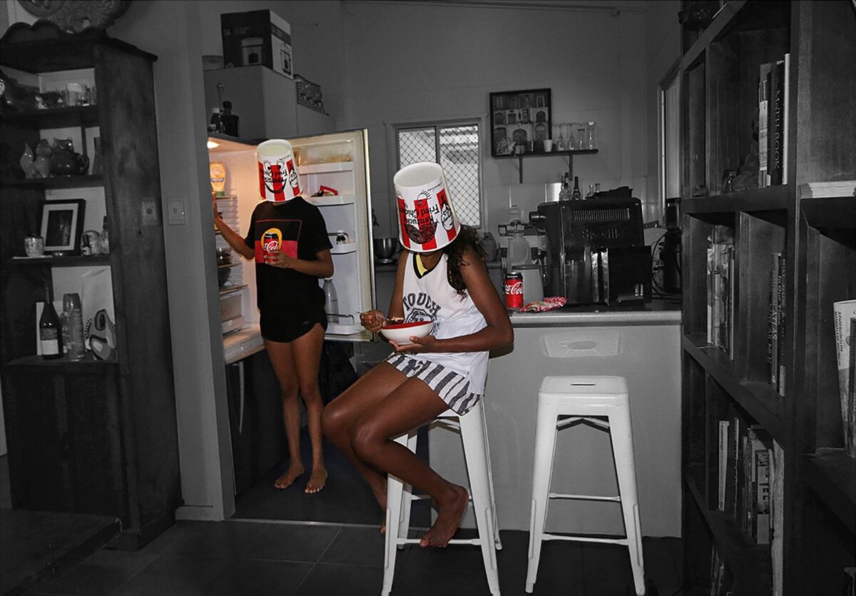 Two young people in a kitchen with empty fried chicken buckets over their heads.