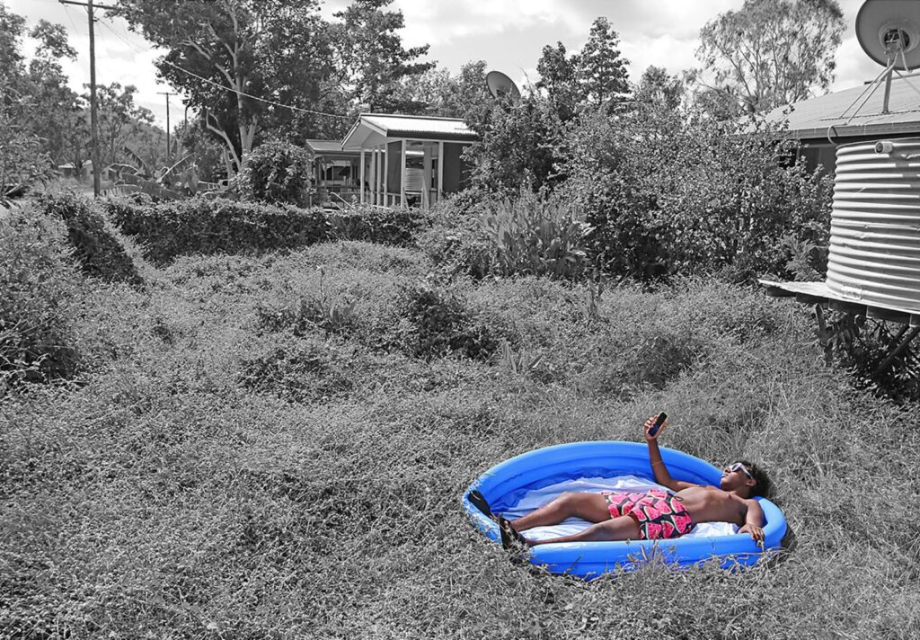 Young person laying in a small, empty inflatable pool in a grassy front yard, looking at their phone.