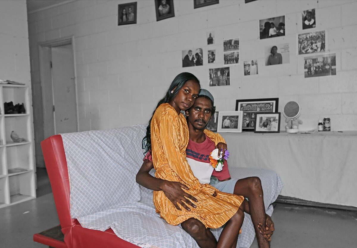 A young couple sitting on a living room couch with photographs on the wall behind them.