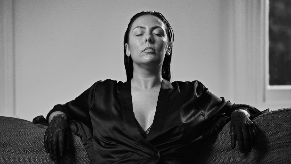 Photograph of a person sitting on a couch with their arms perched upon the back rest. They are wearing a low-cut black silk bathrobe with black gloves on. The figure's head is tilted up, with closed eyes and a peaceful expression.
