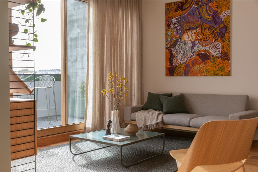 A light-filled lounge room with a couch, chair, glass coffee table, and an abstract painting on the wall, with a curtained floor to ceiling glass door opening onto a balcony.