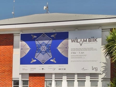 Exhibition banner featuring text and an image in blue and white geometric patterns with birds exhibited on the façade of a brick building.